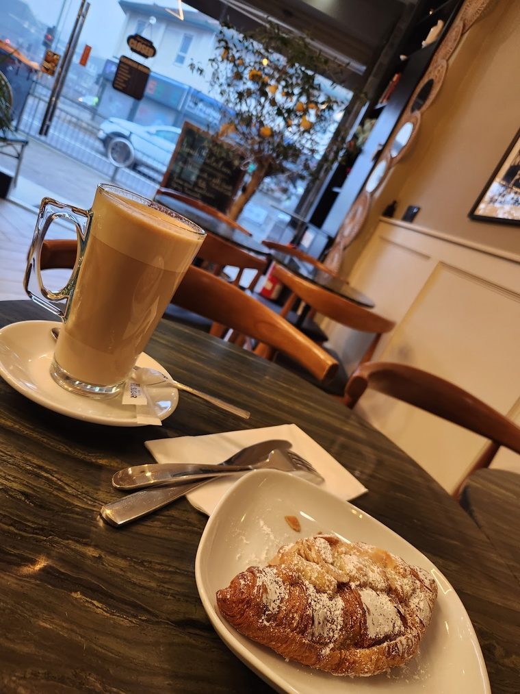 A glass latte beside an almond croissant on the marble window table.
